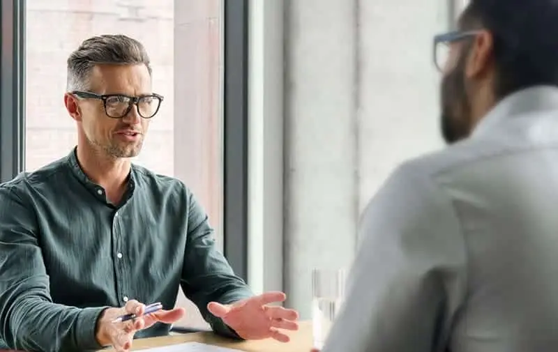 Due uomini sono seduti a un tavolo in una stanza molto illuminata. L'uomo a sinistra, con gli occhiali e una maglietta verde, parla e gesticola con una penna in mano. L'uomo a destra, anche lui con gli occhiali, ascolta attentamente. Sullo sfondo una finestra con vista urbana.
