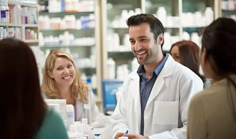 Un gruppo di farmacisti in camice bianco interagisce con i clienti al bancone di una farmacia, circondato da scaffali di farmaci. Un farmacista, in rappresentanza di Evo Sistemi farmacie, sorride e conversa con le persone, mentre gli altri ascoltano e sorridono.